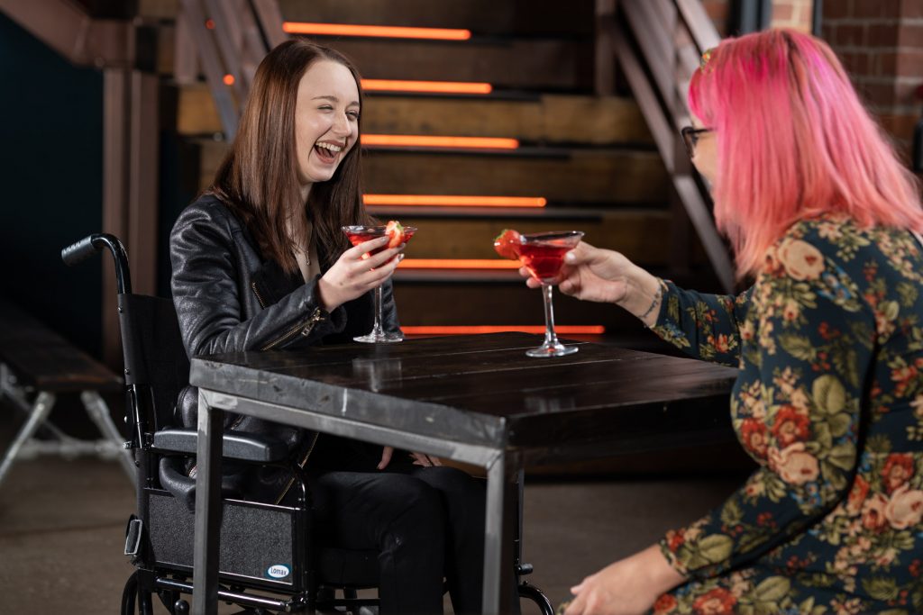 Pippa Stacey and Sam Cleasby are disability bloggers and influencers and sit in a bar drinking cocktails and laughing